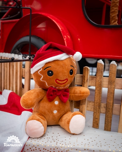 Peluche de bonhomme en pain d'épice avec chapeau de Noël devant une charrette rouge.